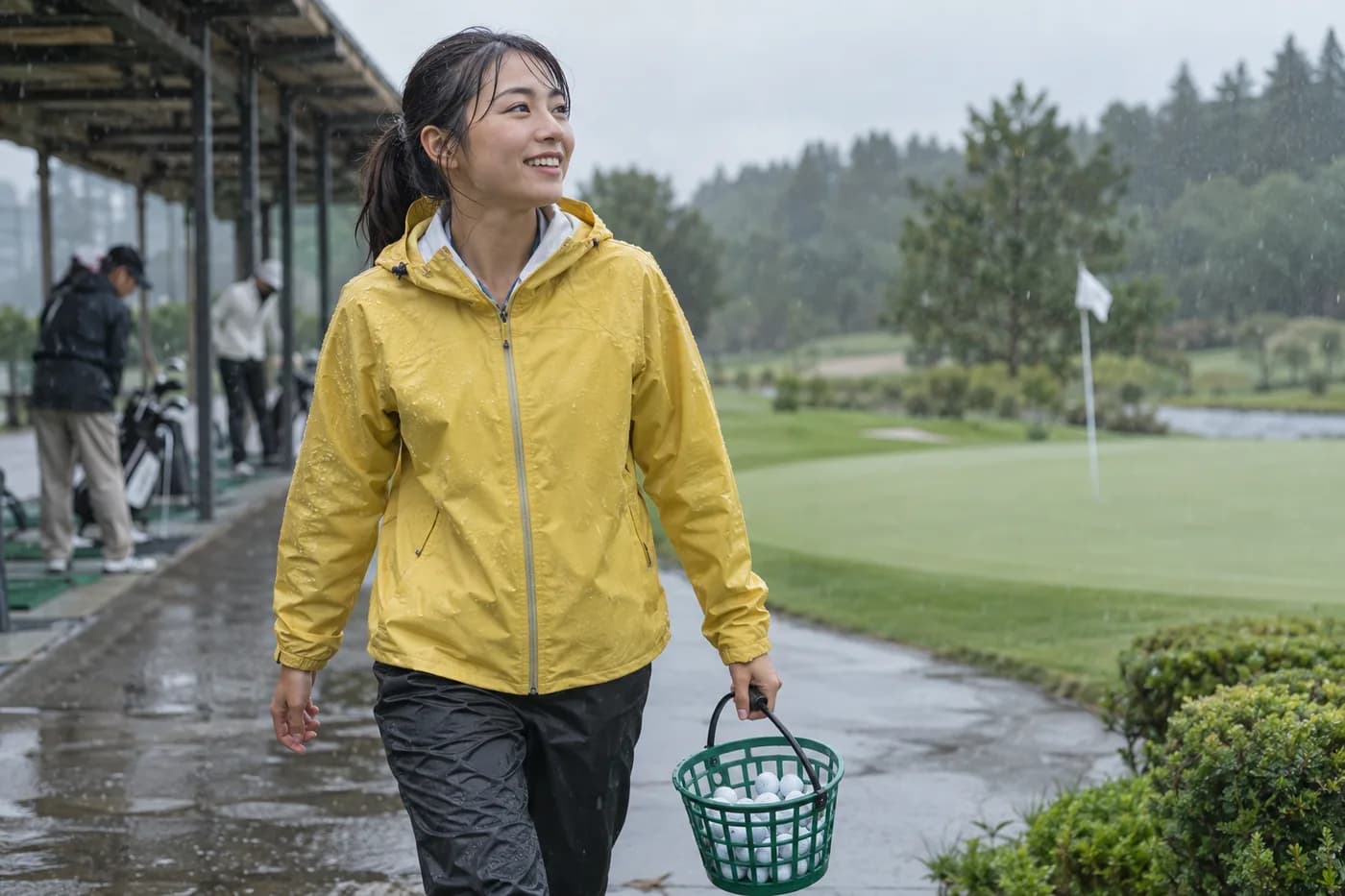 a golfer in a yellow rain jacket carrying a small practice basket from a range toward a putting green