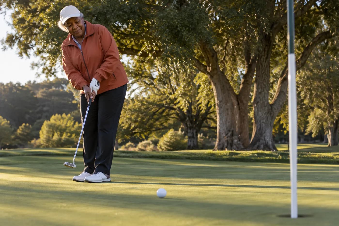 a golfer in a rust-orange windbreaker rolling a putt on a small practice green beside mature trees