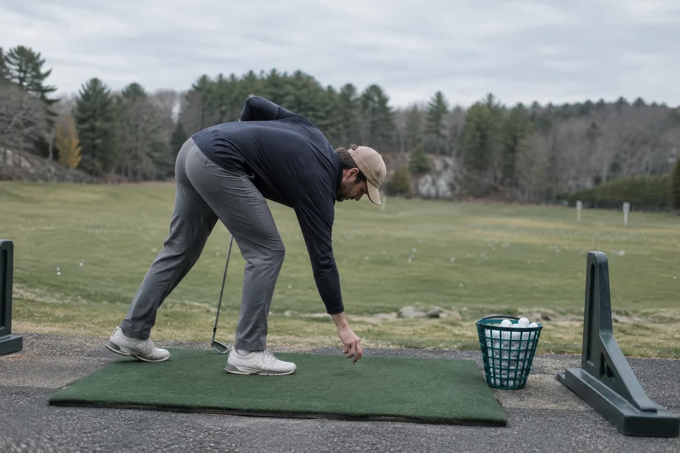a golfer placing a ball on a range mat beside a basket, blurred trees beyond