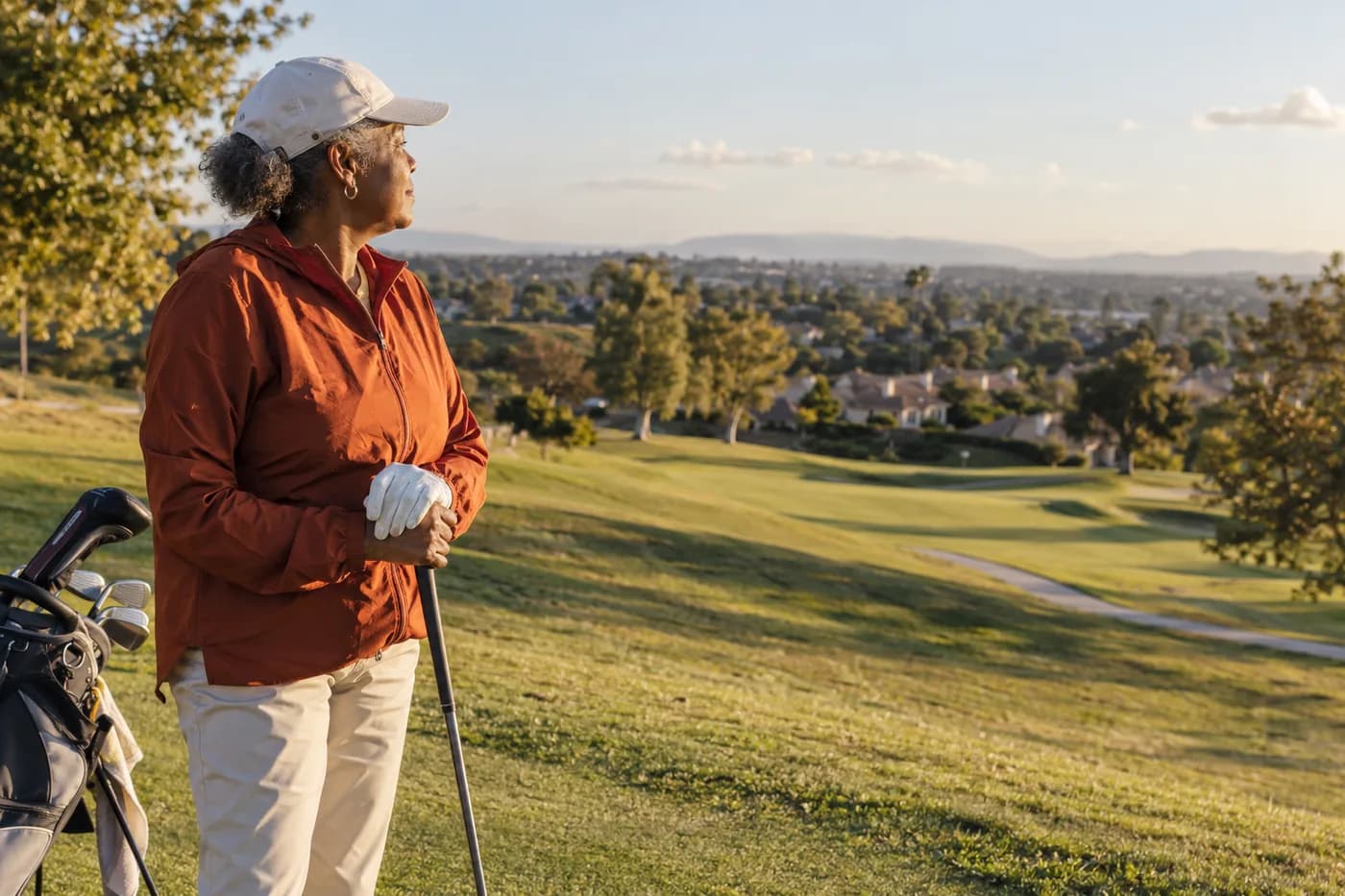 a golfer in a rust-orange windbreaker looking across a sloping neighborhood fairway toward distant rooftops
