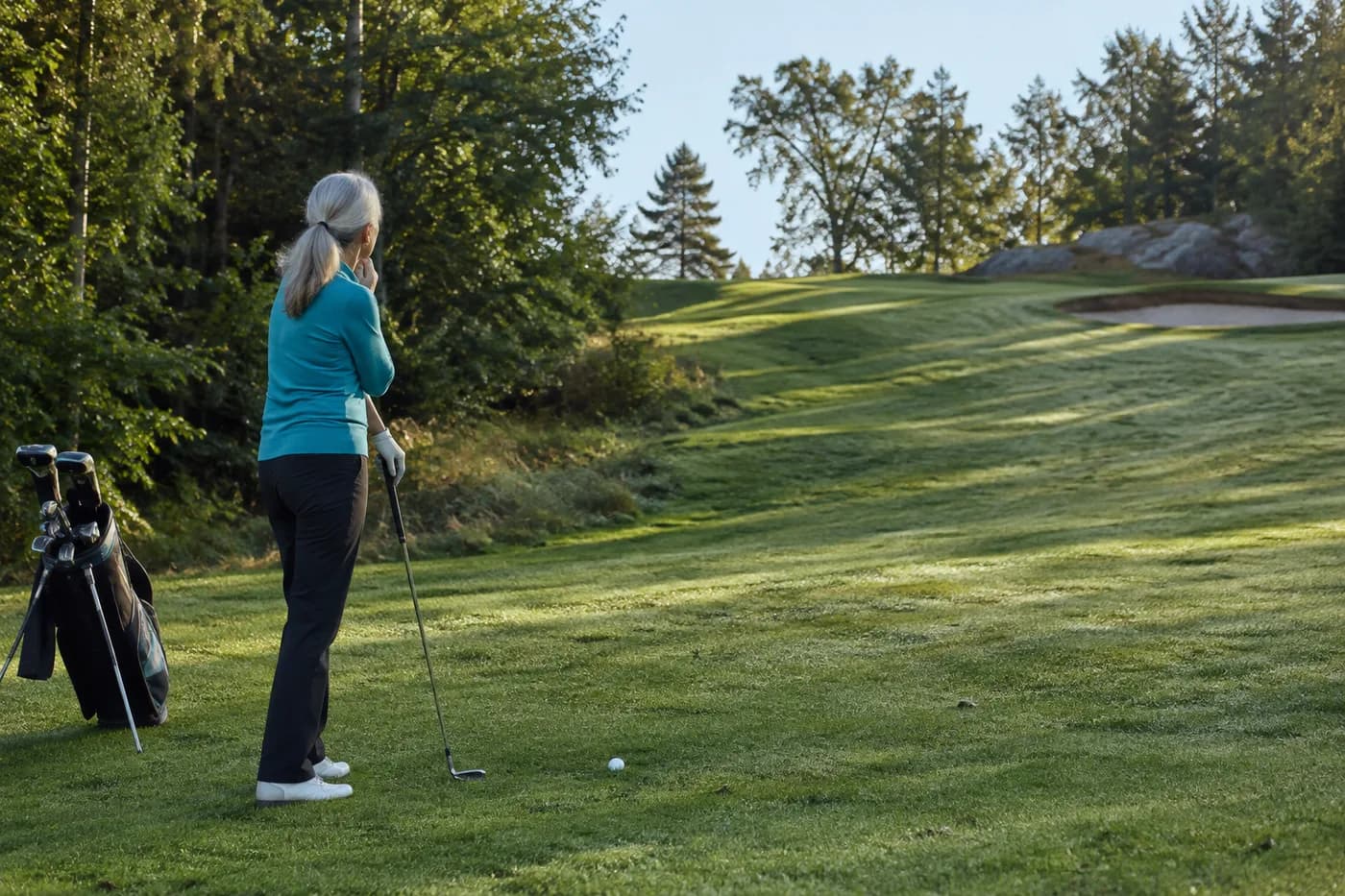 a golfer in a turquoise pullover considering a blind uphill approach beside dense woodland