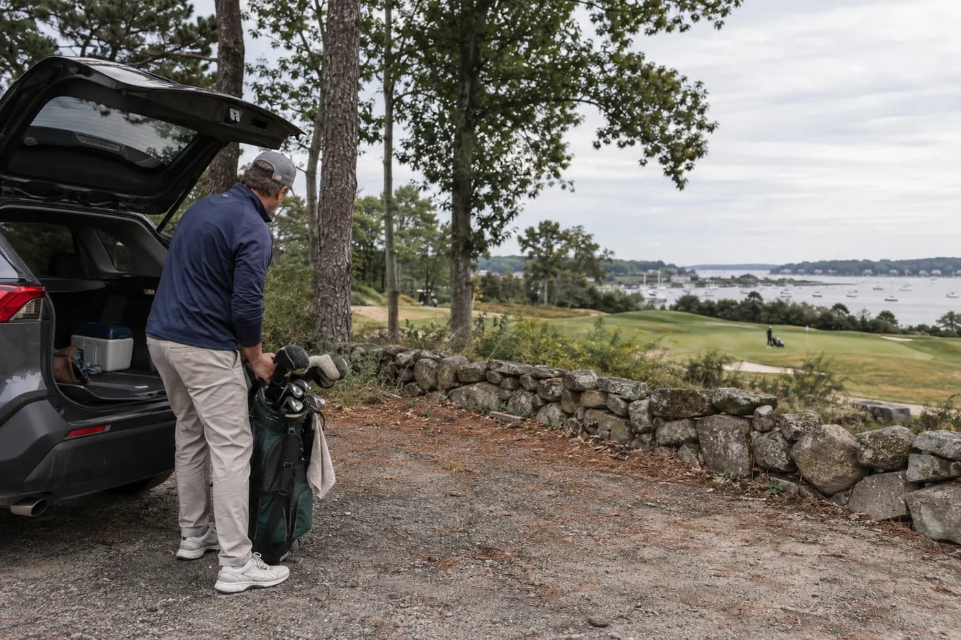 a golfer loading a bag near a wooded lot with a distant harbor glimpse