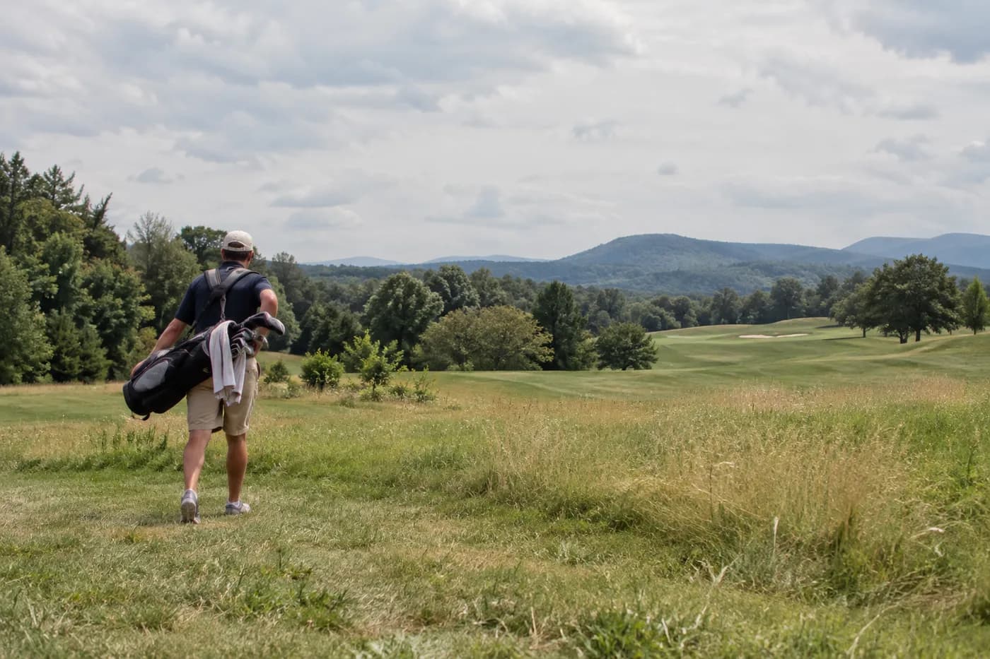a golfer carrying clubs across a broad old farm meadow at midday