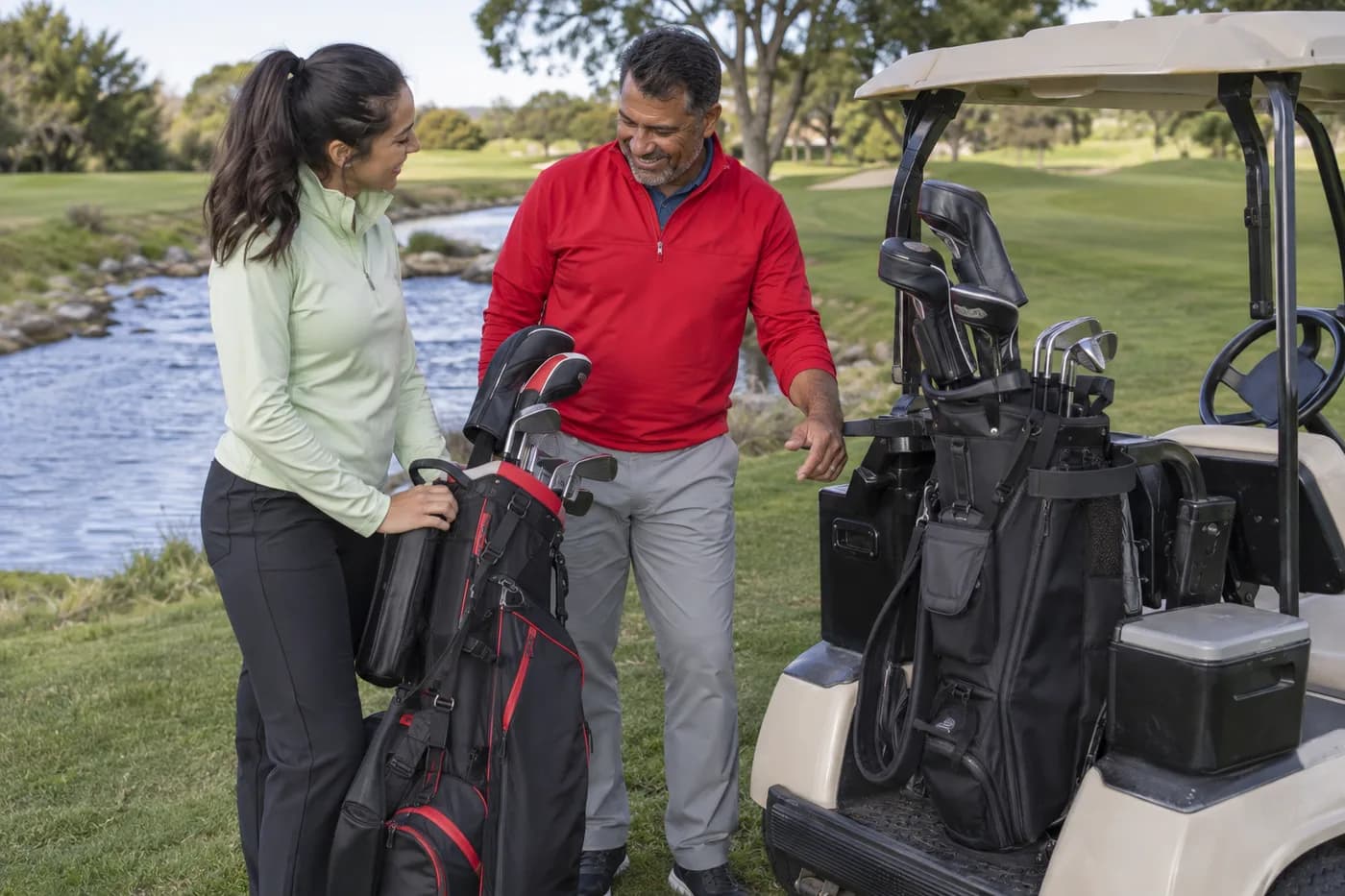 two golfers in red and pale green layers comparing a carry bag and a parked golf cart beside a stream