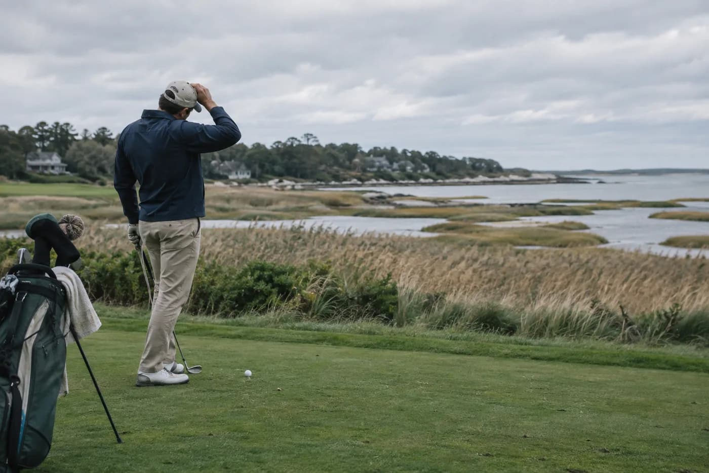 a golfer holding a hat against coastal wind beside saltmarsh reeds