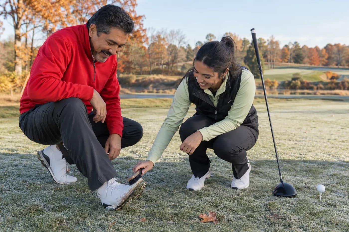 two golfers in red and pale green layers brushing frost from a shoe next to an autumn leaf on a tee box