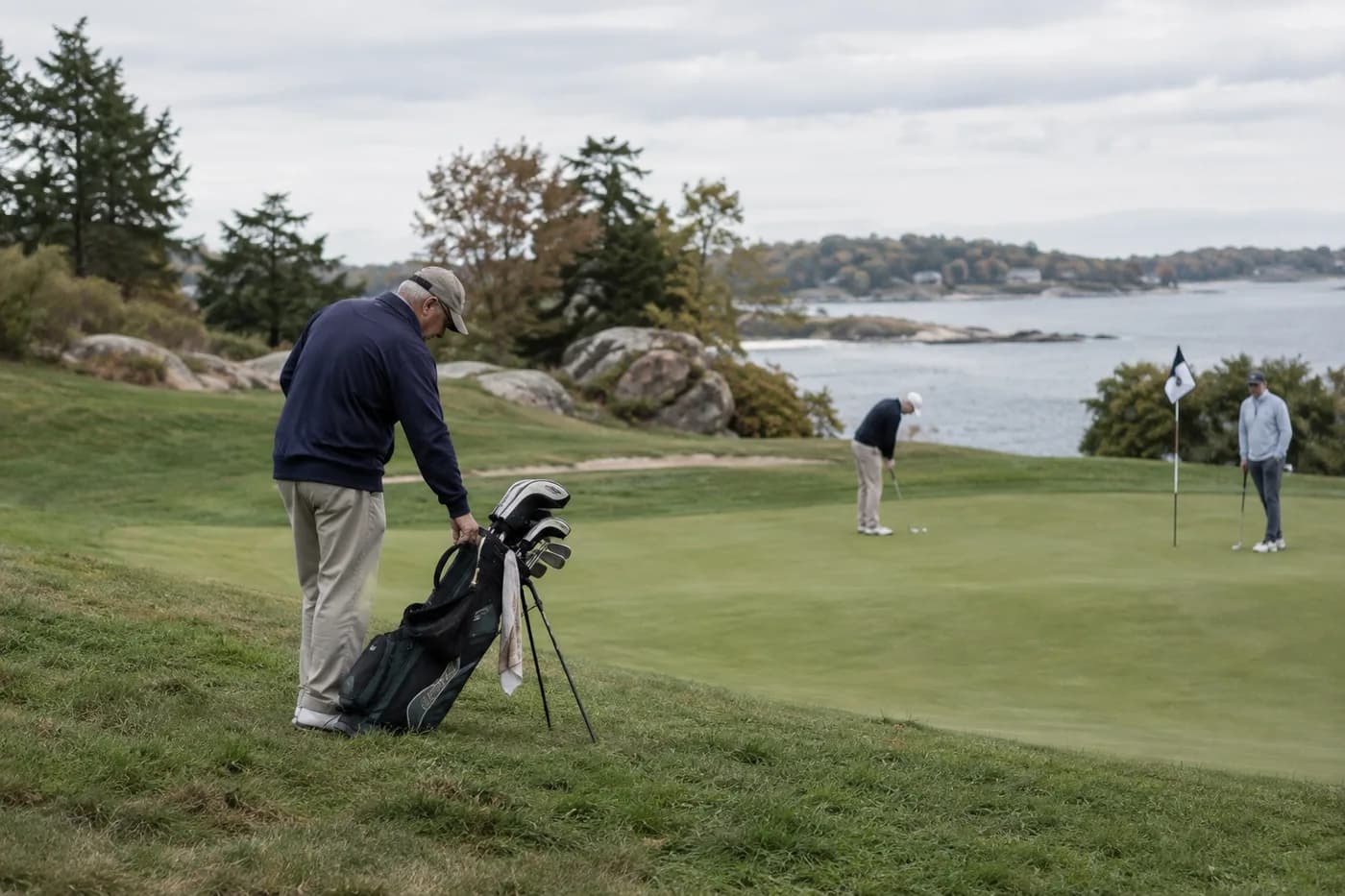 a golfer steadying a bag on a rolling hill beside a putting green