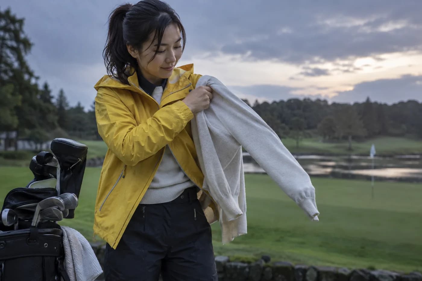 a golfer in a yellow rain jacket putting on a light sweater beside a carry bag as the evening cools